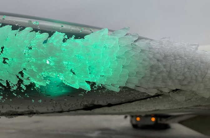 Ice crystals formed along the edge of an airplane wing. Some crystals appear to glow green as they cover a light on the wing.