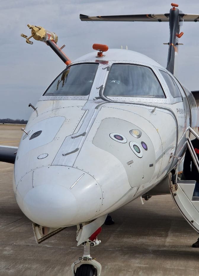 The optical sensor looks like a glass circle embedded in the nose of a business jet. The microwave sensor is near the optical sensor, and it looks like a flat golden ring, embedded in a nearly flat, tear-drop-shaped case. Two other similar looking sensors from other research groups are installed nearby. The scientific instrument is mounted on a pipe protruding above the cockpit.