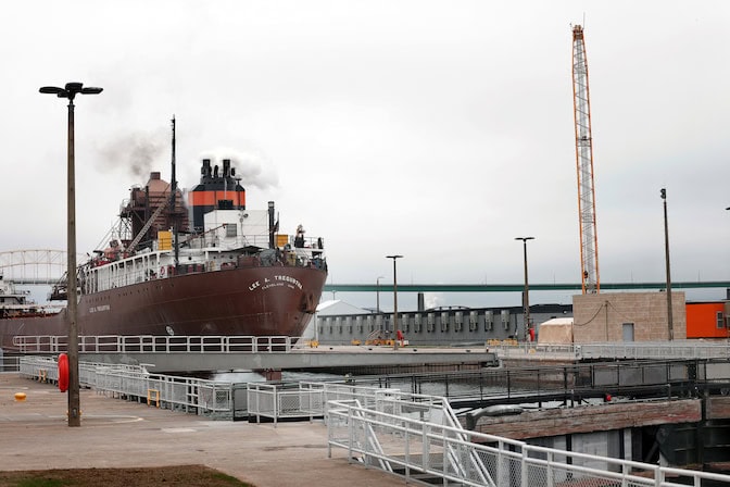 A view of the stern of a large commercial ship as it leaves the locks. It towers over the walkways that surround the reservoir the ship is exiting. The ship's name is printed in white on the hull.