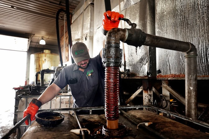 A man opens a valve on a pipe connected to an orange hose. The hose extends from the pipe into a plastic tank, which is covered with black grime