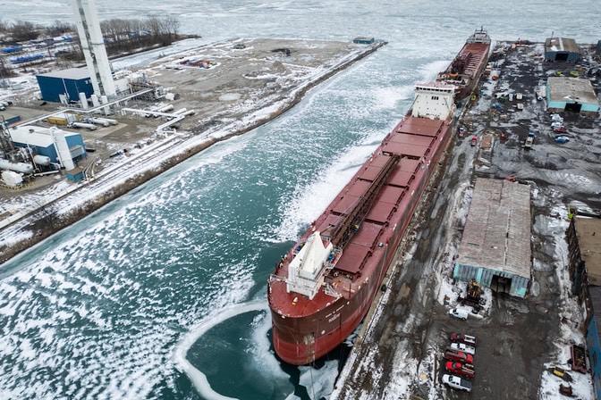 Two massive ships moored at a dock. The freighters dwarf the nearby storage garages. Snow piles dot the dock and the nearby water is frozen.