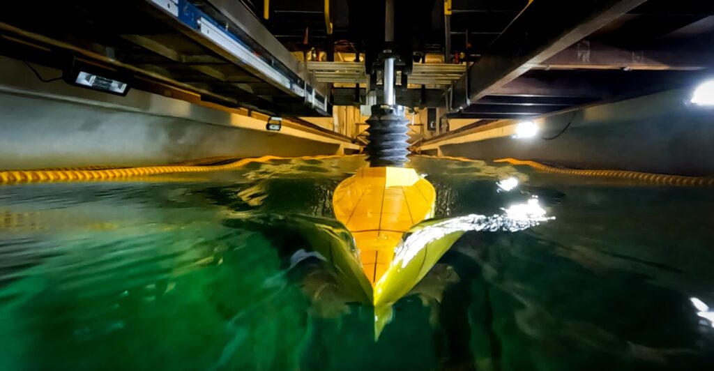 The bow of a model ship, which resembles a thin, yellow wedge, points directly at the viewer. Its deck is only a few inches above the waterline. A wave near the front of the ship swells higher than the ship's deck near the tip of the bow.