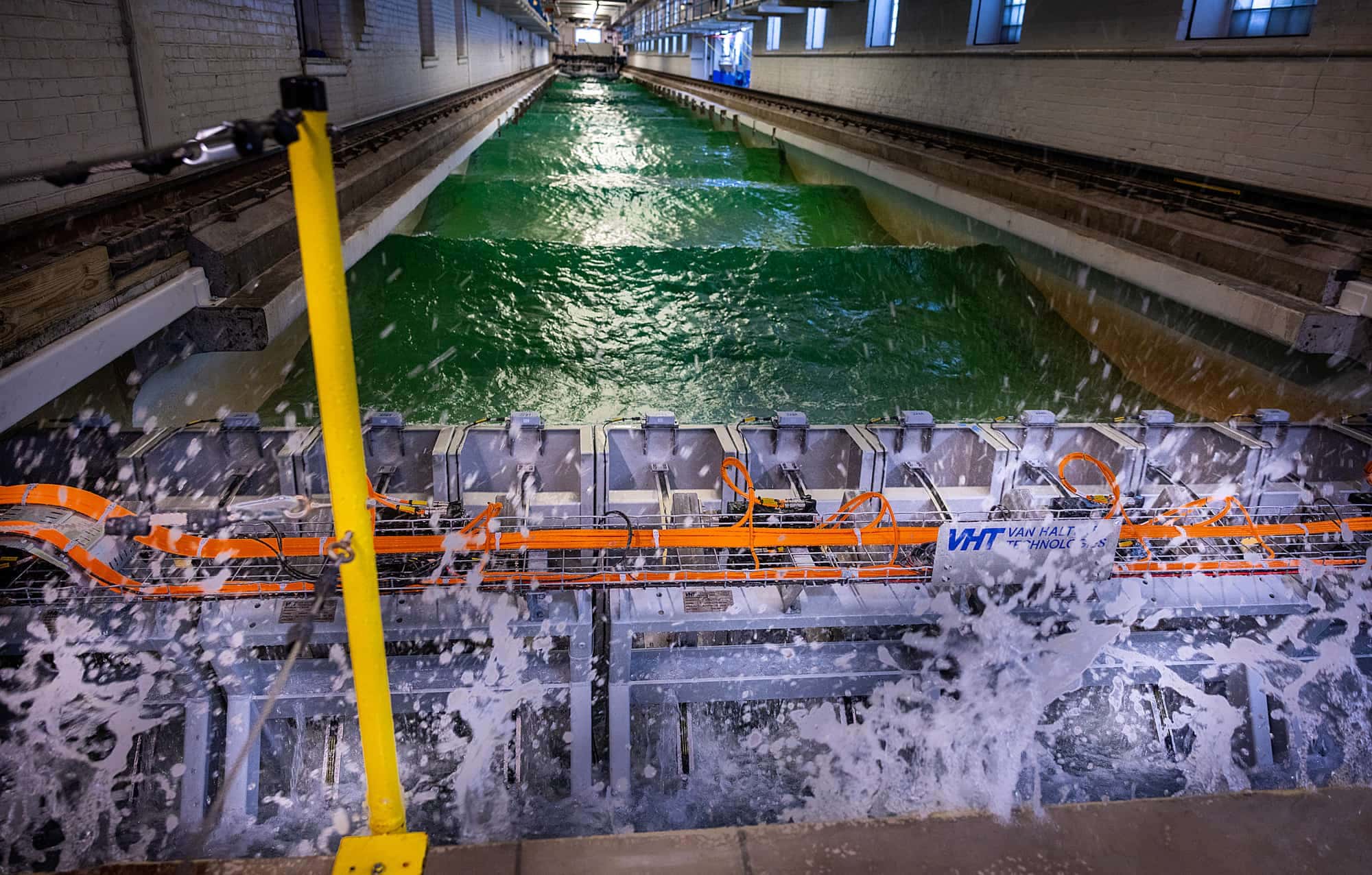Water splashes around a set of steel panels at the edge of the tow tank. Waves crest down the length of the tank.