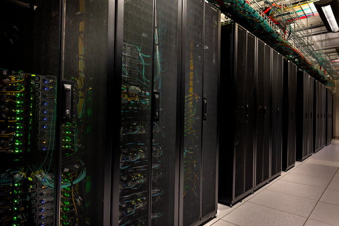 Black server towers stretch down the length of a hallway in a tightly packed row. Each tower contains stacks of many computers. Green lights blink through the grated, black doors that secure each tower.