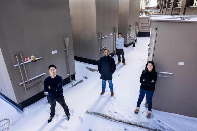 Three men and a woman stand in a triangle formation among generators outside a computing center. The generators are inside tan sheds that are two or three times taller than the researchers. There is snow on the ground.