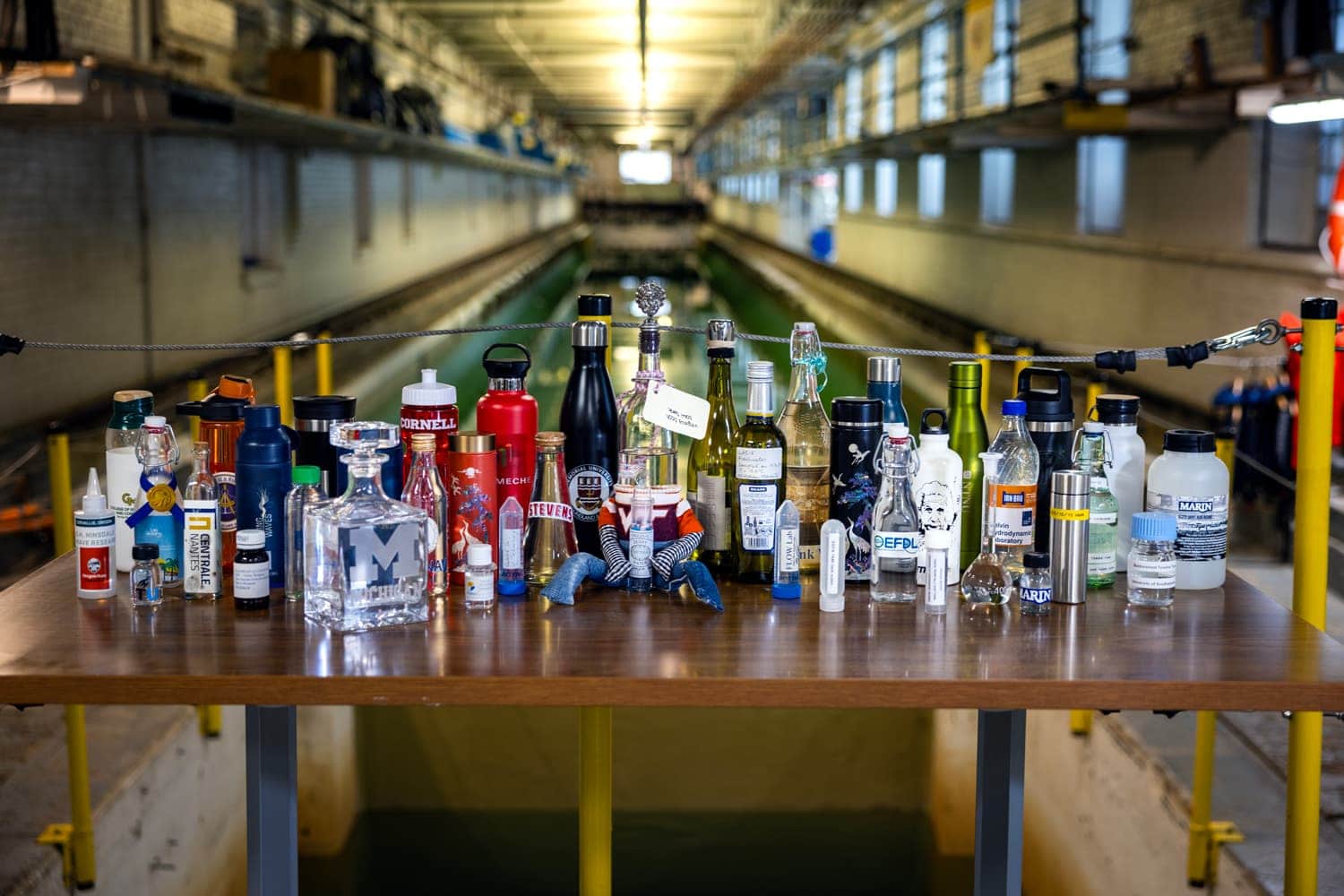 A collection of bottles and glassware are arranged on a table in front of a long tank of water. The logo or name of a lab is printed on each bottle.