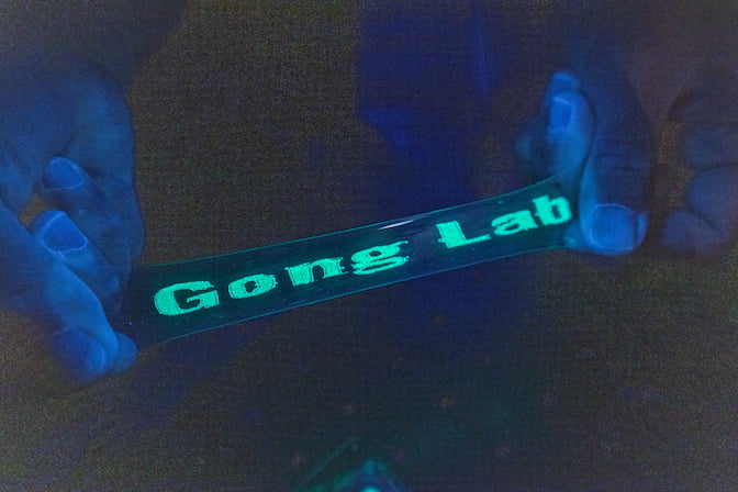 A transparent film is stretched between two hands. A blacklight reveals a message on the film, which reads "Gong Lab" in neon green lights.