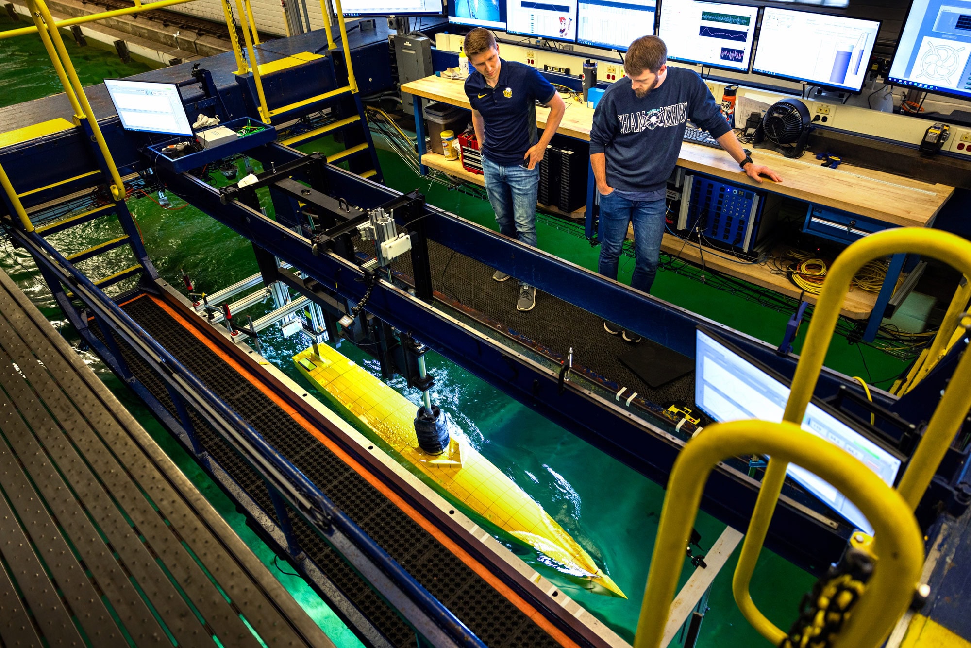 Engineers observe a yellow scale model vessel being tested in a tow tank, with sensors and monitoring equipment mounted above the water and data screens in the background.