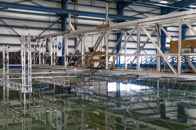 A shallow, concrete pool inside of a warehouse. Metal scaffolds about a meter above the water. Two people sit at desks looking at their computer on the scaffold.