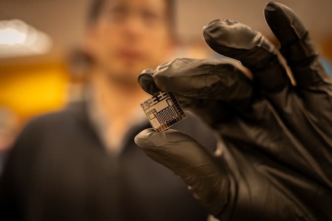 A man blurred in the background holds an in-focus, square chip, about the size of the tip of a finger, between his index finger and thumb while wearing black latex gloves. The black and gold chip contains three T-shaped entrances at the top edge and a golden rectangle and serpentine coil on the bottom edge.