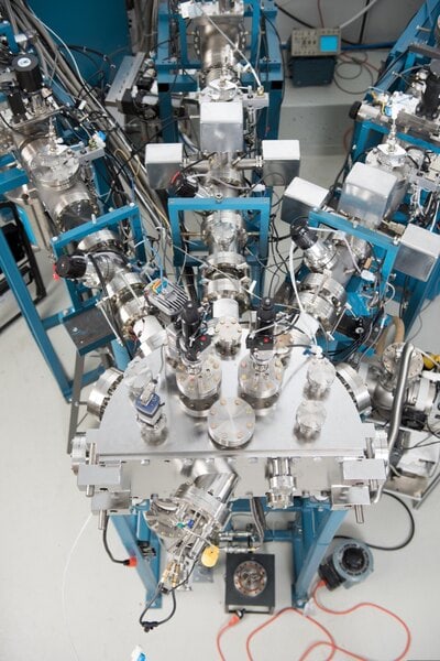 Three complicated-looking beam lines, composed of silvery cylinders and boxes, converge on a half-moon-shaped target box, itself covered in ports. Many wires protrude from the system.