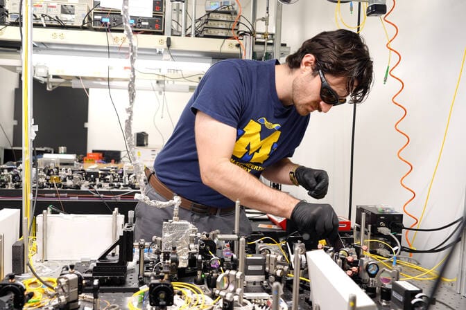 A man in black gloves and laser safety glasses stands at a crowded optical table with various elements attached, some with wires running to them.