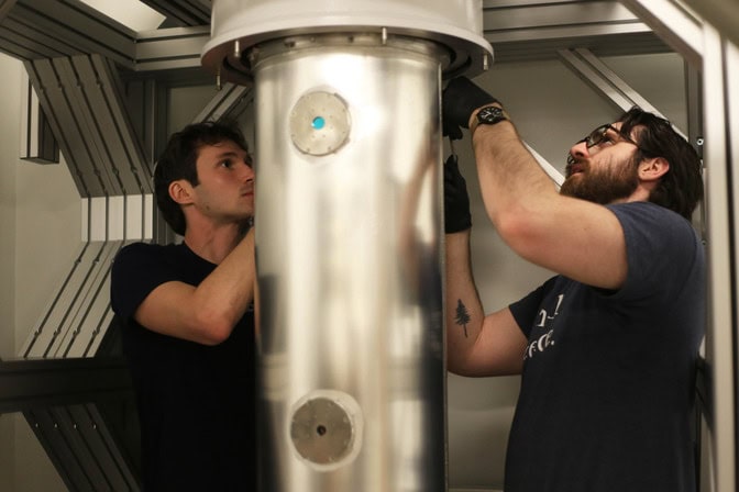 The dilution refrigerator looks like a white cylinder about two feet in diameter with a slightly narrower silver-tone cylinder beneath. Amozeg and Ward, wearing black gloves, are loosening the attachment points between the cylinders.