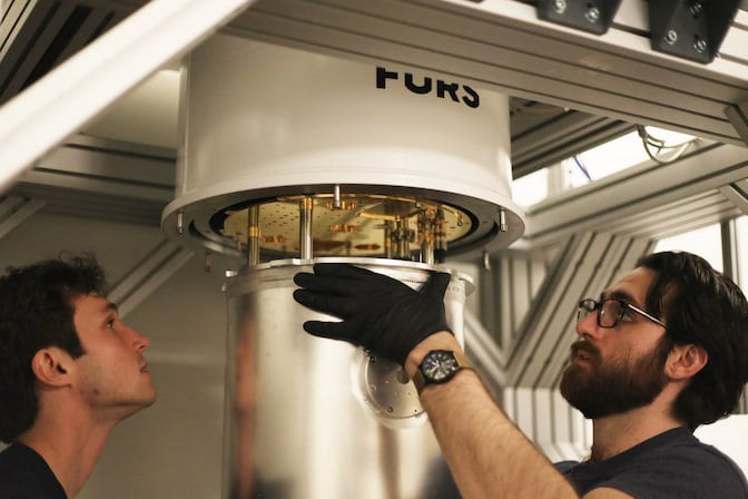 The dilution refrigerator looks like a white cylinder about two feet in diameter with a silver-tone cylinder beneath. Ward, wearing black gloves, has lowered the silver cylinder by a few inches, revealing a golden disc with silver rods extending downward.