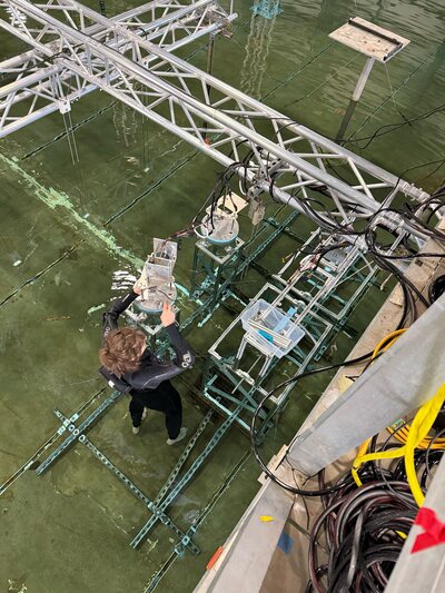 A person in a wetsuit stands in ankle deep water in a concrete pool. They hold a wrench in one hand while steadying a circular metal platform with the other hand, tightening screws. The platform is connected to another box with wires extending to an out-of-site source on the edge of the pool.