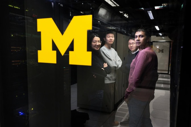 A yellow University of Michigan "M" logo is in the foreground in a server room as four people in the back look at the camera near the servers.