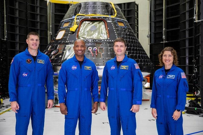 Four astronauts stand in front of a cone-shaped spacecraft, which is parked in a large lab space.