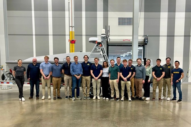 A group of students, engineers, and professors stand in front of a vessel on a trailer that is parked indoors.