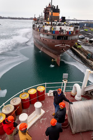 A group of engineers stand on the deck of a large commercial ship moored at a dock. Another large commercial ship is stationed nearby.