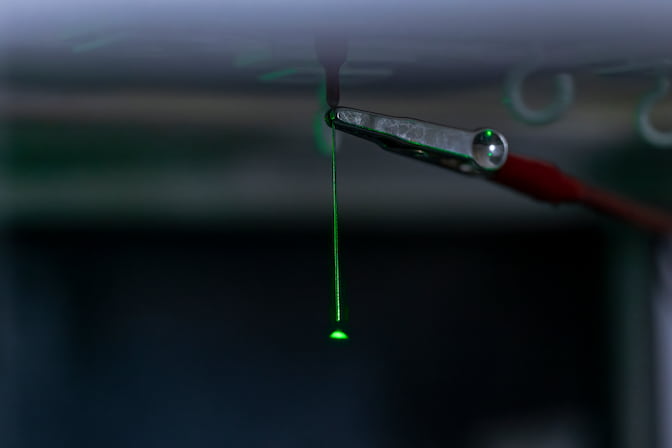 A tiny, green cone of light fans out from the tip of a syringe. An electrode clamp is attached near the base of the needle.