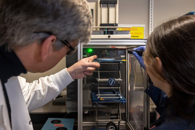 A woman shines a green laser into a transparent box, aiming just below a syringe that extends into the box through a hole in the top. A man points at the syringe inside the box. A jack stand holds the aluminum plate a few inches beneath the syringe needle.