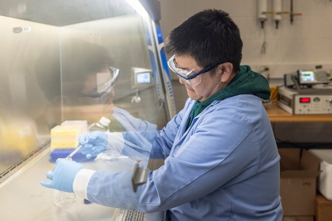 A graduate student wearing a lab coat, gloves, and protective glasses uses a dropper to add clear liquid to a test tube.