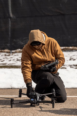 A person wearing a tan coat and gloves kneels on the ground to set up a drone for flight.