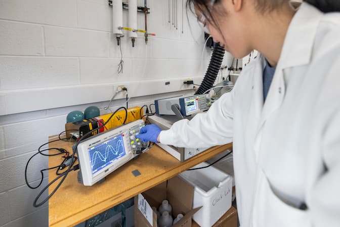 A graduate student wearing a lab coat and gloves turns the knob on a box-like machine, called an oscilloscope, that has a small screen.