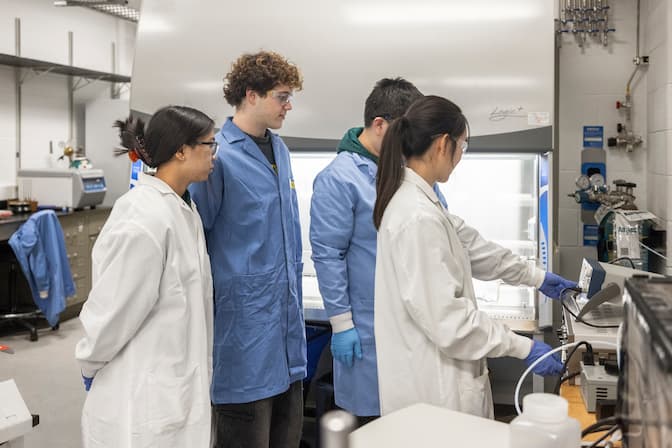Four students wearing lab coats, gloves, and protective glasses stand in a lab space. One of the students points at a piece of lab equipment in front of them.