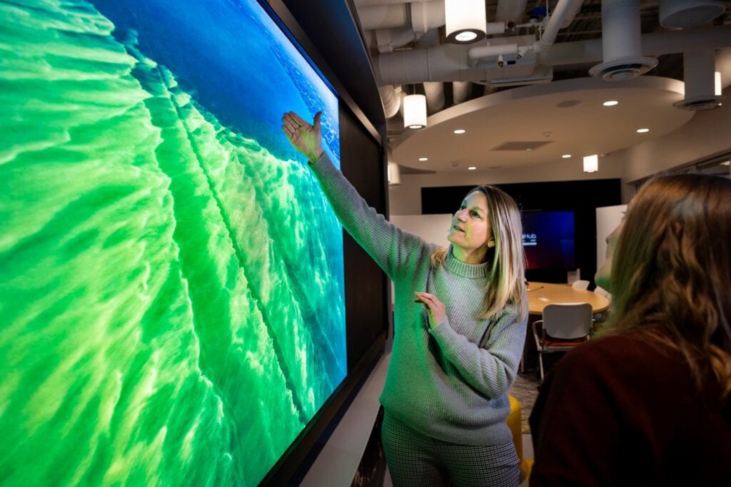 Woman gesturing with both arms toward a large video screen showing an algal bloom