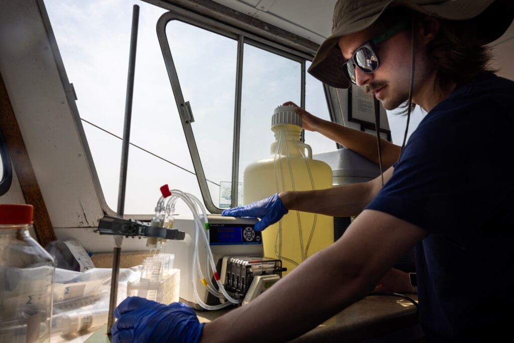 Man in a bucket hat and sunglasses works with chemistry equipment on a benchtop situated  next to an open boat window