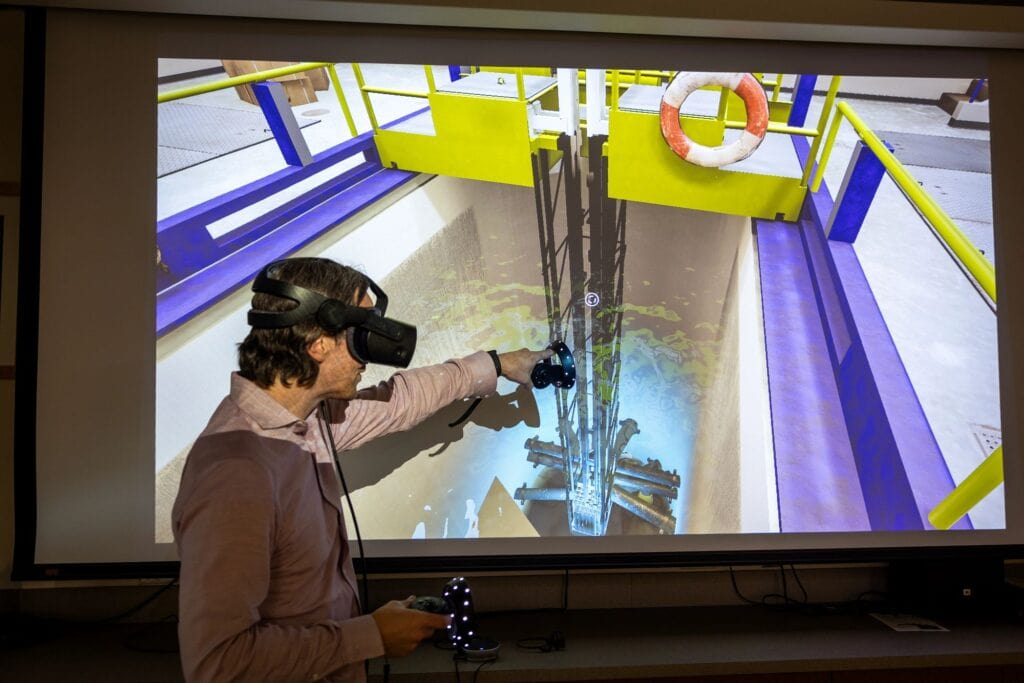 Man wearing a VR headset points a controller at a large projection of a virtual reactor core in a lab setting.