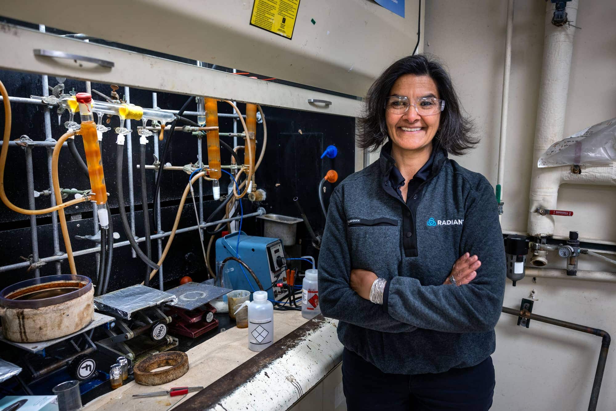 The woman stands in a lab with arms crossed, smiling in front of scientific equipment, tubing, and instruments.