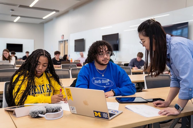 Industrial and Operations Engineering students collaborate during class at the University of Michigan, with one student taking notes, another working on a laptop with U-M IOE stickers, and an instructor leaning in to assist at their table in a modern classroom.