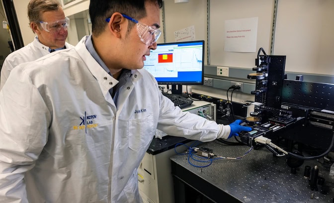 Two men wearing lab coats stand in front of a device that looks like a series of stacked lenses. One man places a piece of plastic-like material beneath the lenses. A nearby computer monitor displays a series of graphs.