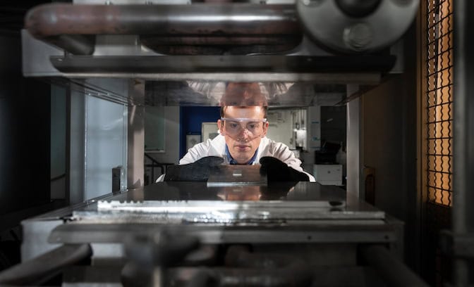  A man slides a metal tray between the plates of a large hot press. A rectangular slide loaded with silk fibers rests on the tray.