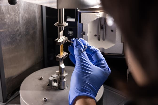 Metal clamps latch onto a thin, rectangular piece of amber-colored plastic-like material at its top and bottom edges. A man uses an Allen wrench to adjust the top clamp. A kiln-like chamber is open behind the clamps.