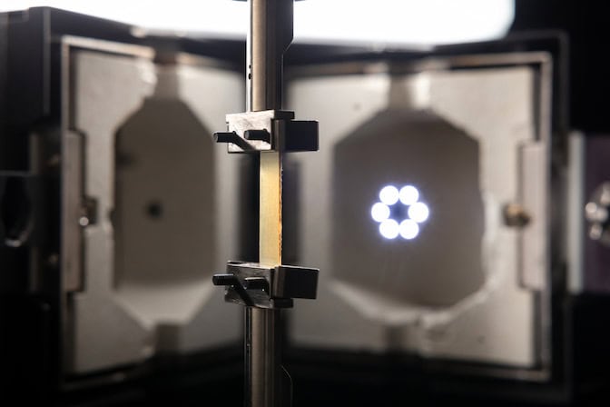 Metal clamps latch onto a thin, rectangular piece of amber-colored plastic-like material at its top and bottom edges. A kiln-like chamber is open behind the clamps.