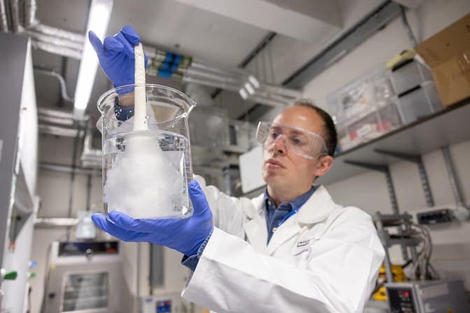 A man wearing a white lab coat, nitrile gloves and safety goggles holds a glass beaker in one hand. The beaker is filled with water and long silk threads, some of which the man is pulling out of the water with his other hand.