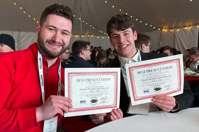 Justin Hamil (left) and Alexander Birmingham (right) sitting at a table in a tent, smiling and each holding a certificate that reads “Best Presentaiton.”
