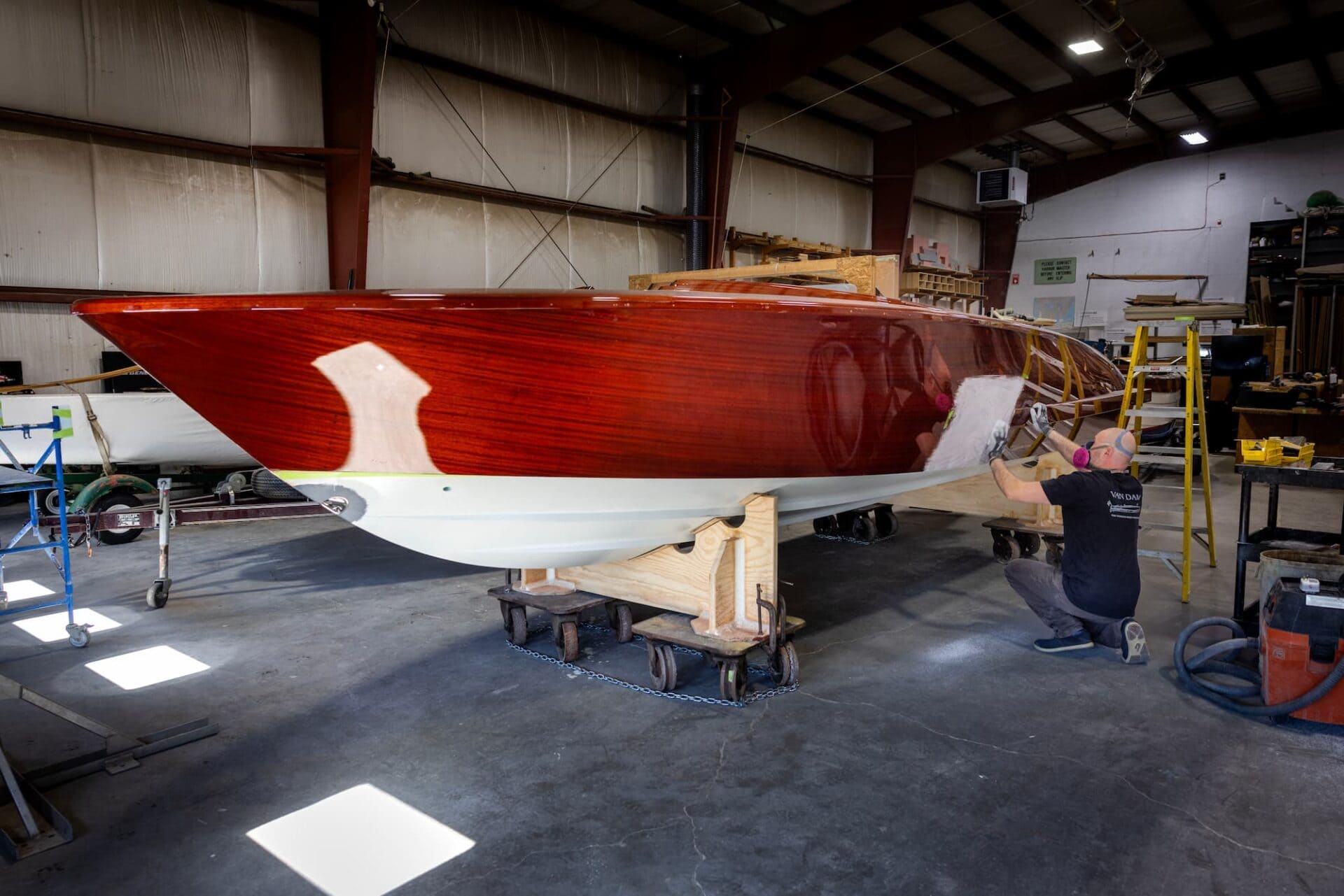 A worker applies a finish to a wooden boat in a workshop. The boat has a glossy mahogany upper and white hull, supported on a wheeled cradle.