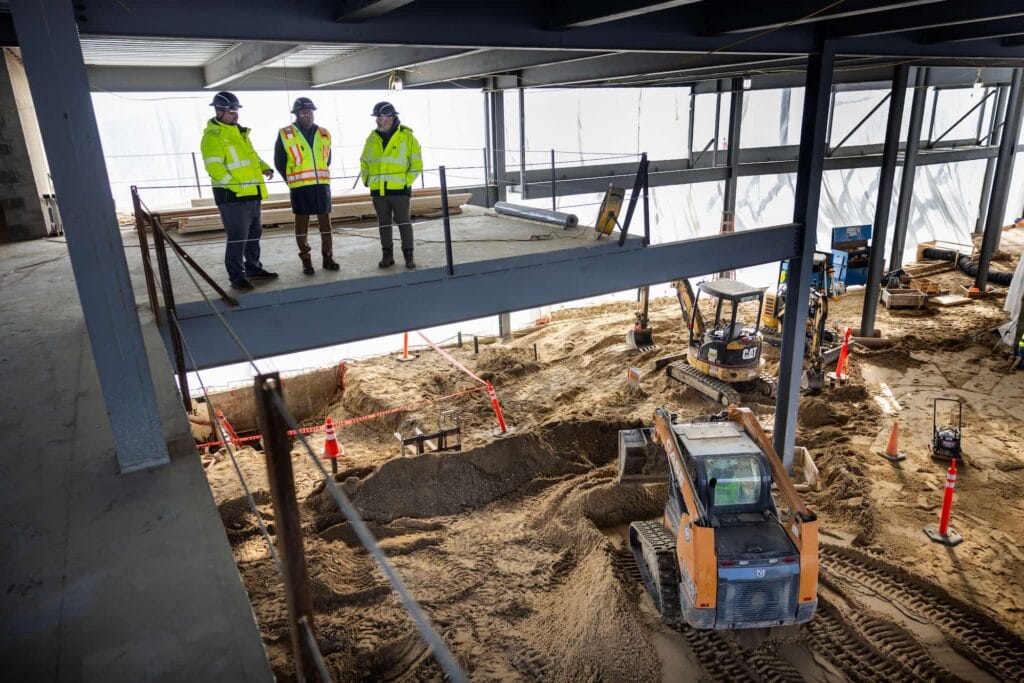 Three men on a platform watch a construction site with men in hard hats working and driving heavy equipment.