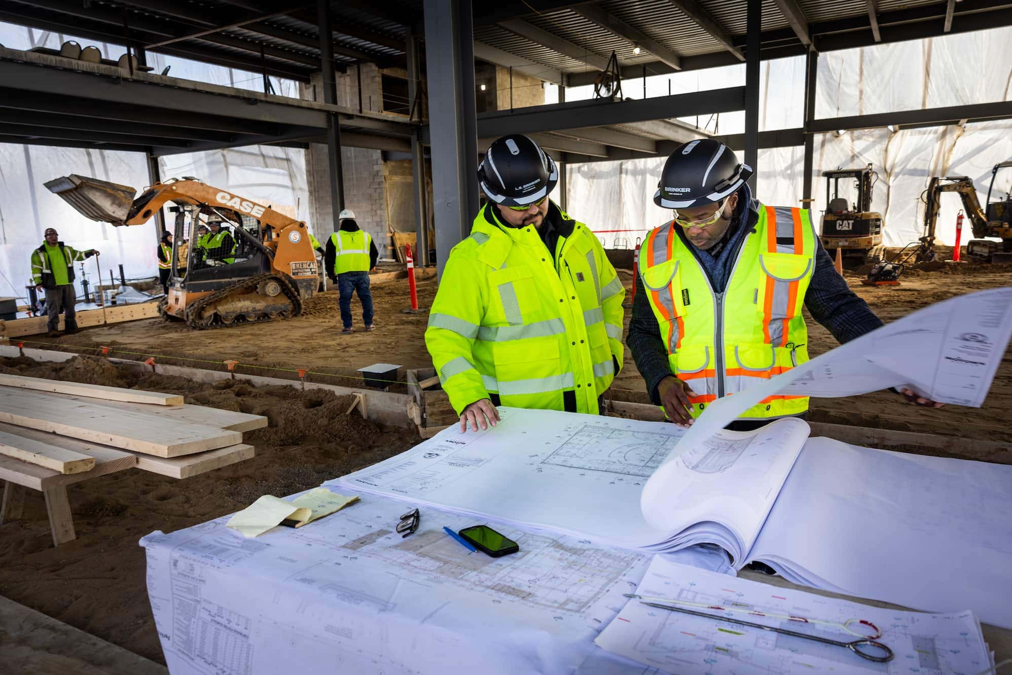 Two men consult blueprints on a construction site with men in hard hats working and driving heavy equipment behind them.