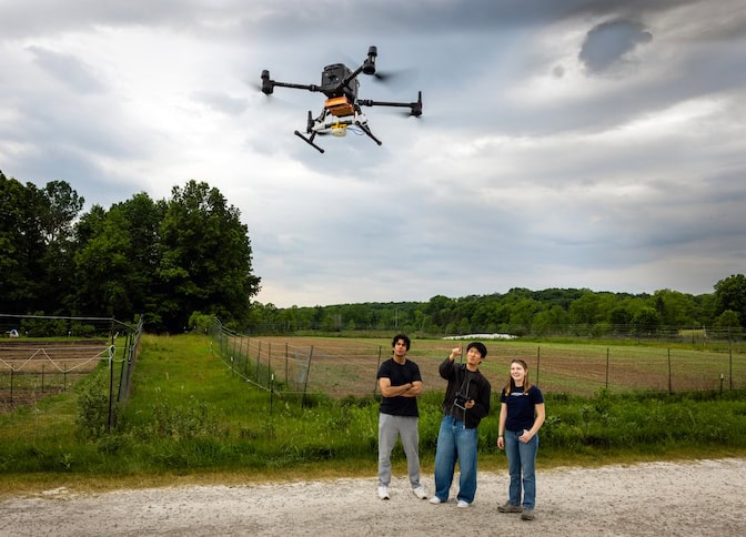 A drone is in the air over the heads of three students, who are standing near a farm field. The drone has four arms with propellers at each end.