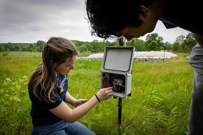 A woman knees to adjust electronics inside an open gray box. The box is hanging from a post stuck into the ground. Another student leans forward to see inside the box.