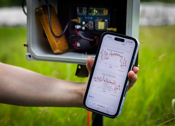 Two line graphs are displayed on a phone held in someone's hand. The graphs have jagged red lines. The phone is held in front of a gray box with electronics inside.
