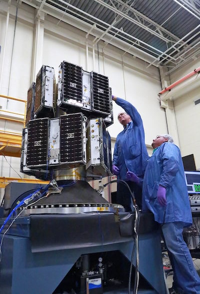 Eight trapezoid shaped satellites are attached to a metal post in two rings of four. Two engineers wearing blue lab coats and hair nets inspect the satellites.