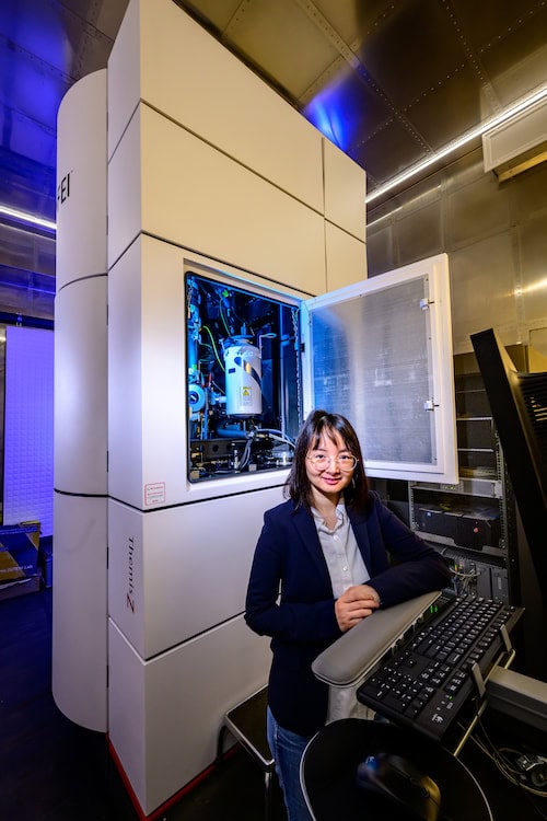 A woman stands at a desk, her arms folded on a keyboard. Behind her is a large instrument that resembles a cabinet. A door on the instrument is opened, revealing many wires and a cylinder-shaped device.