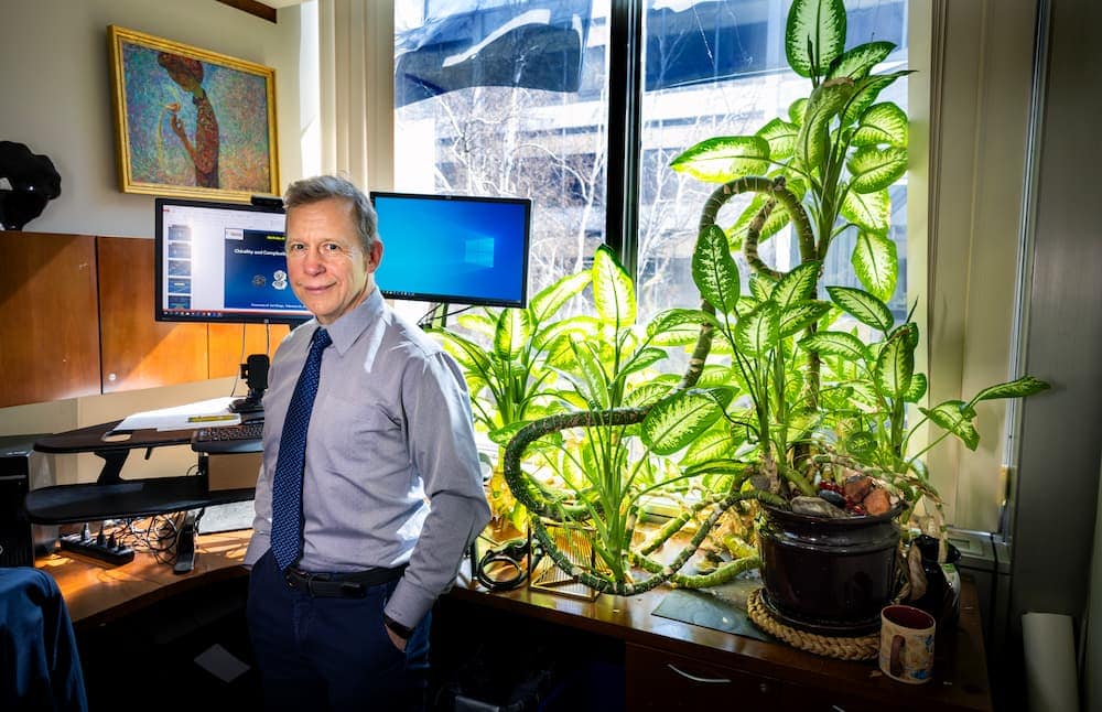 A man stands in an office. His desk is behind him. The thick stem of a plant coils around his desk. The plant's large variegated leaves are lit by sunlight streaming through a large window behind the desk.