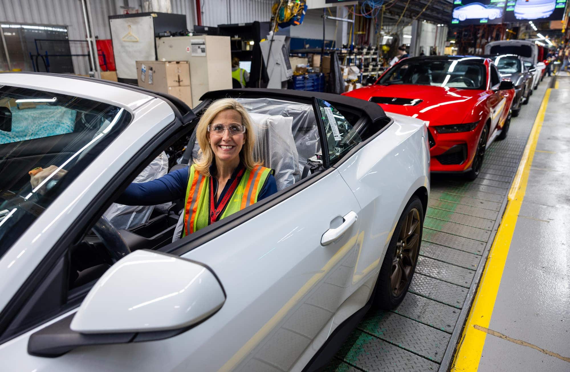 Laurie Transou wearing a safety vest sitting in a white Ford Mustang convertible on the assembly line, smiling as finished cars line up behind her.
